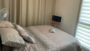 Ein Schlafzimmer mit Bett, beigen Vorhängen, einem Standventilator, einem kleinen Tisch mit Bildschirm und einem Fenster mit Jalousien.
