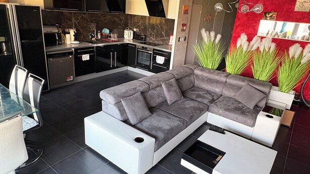 Ein modernes Wohnzimmer mit einem grauen Ecksofa, einem Essbereich mit einem Glastisch und einer Küche mit dunklen Küchenschränken.