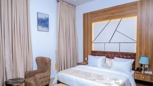 A hotel room with a large bed, a wooden headboard, a side table, a chair, and a small table. - Grovehill Hotel Plus Ilesa  (Ilesa)