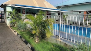 A swimming pool with a metal fence, surrounded by greenery and a building with a blue roof. - Grovehill Hotel Plus Ilesa  (Ilesa)