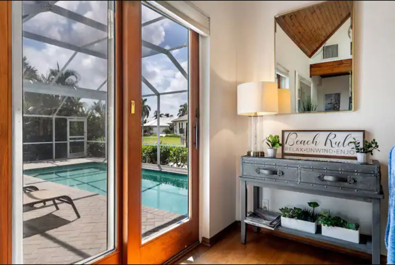 A room with a large sliding glass door leading to a pool area, a console table with a lamp, and a mirror with a 'Beach Rules' sign.