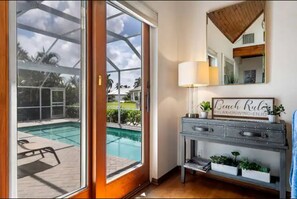 A room with a large sliding glass door leading to a pool area, a console table with a lamp, and a mirror with a 'Beach Rules' sign. - New!Waterfront Pool+Cabana+GameRoom AmazingViews (Marco Island)