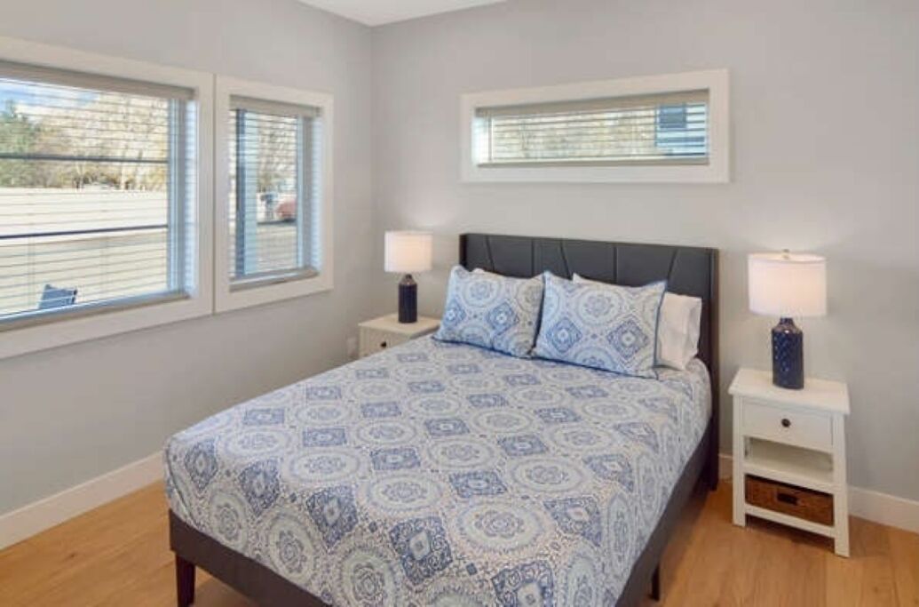 A bedroom with a bed, two bedside tables, and two windows.