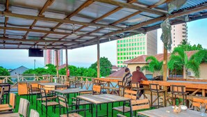 An outdoor dining area with wooden tables and chairs, a covered roof, and a view of a tall building in the background. - K27 Premier Backpackers (Dar es Salaam)