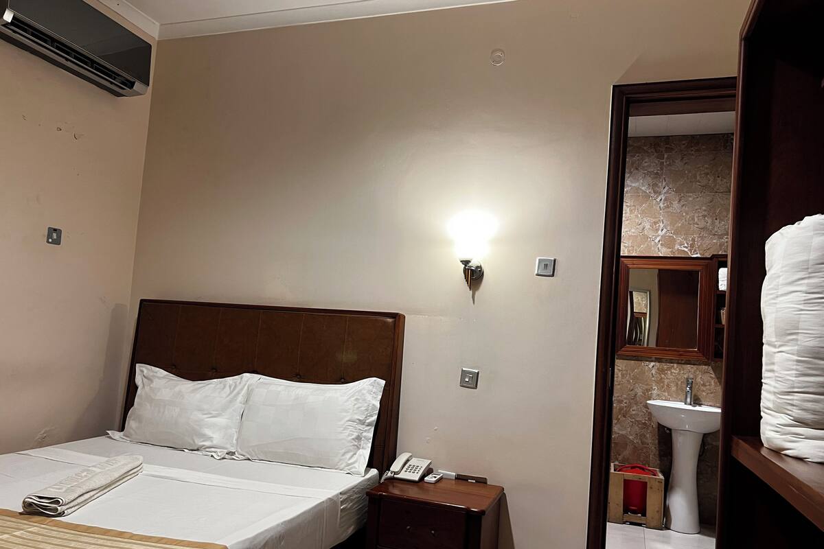 A hotel room with a bed, bedside table, lamp, and a bathroom visible through an open door.