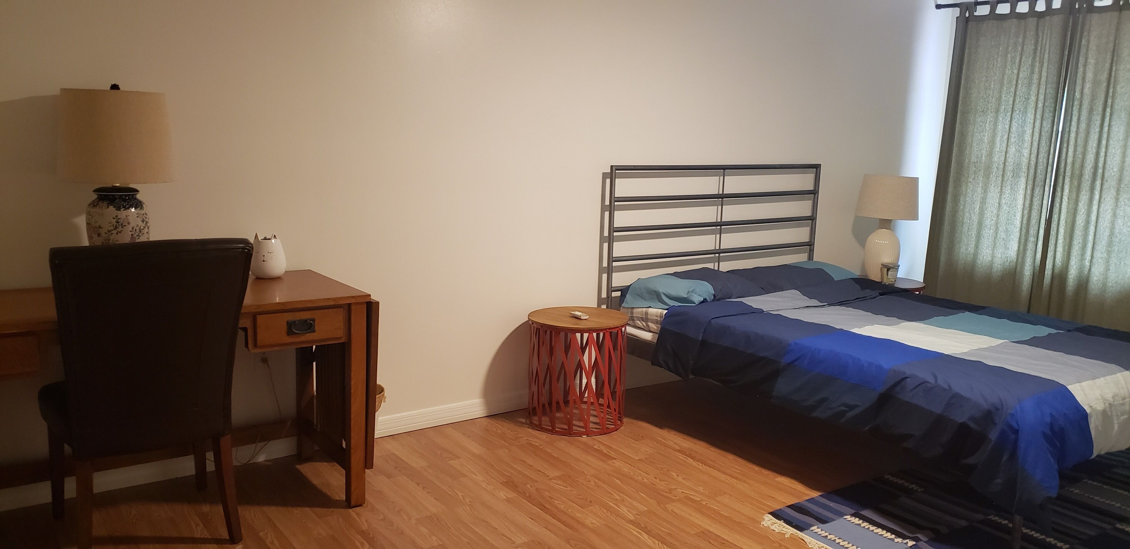 Een slaapkamer met een bed, een bureau, een stoel, een klein rond tafeltje en een lamp.