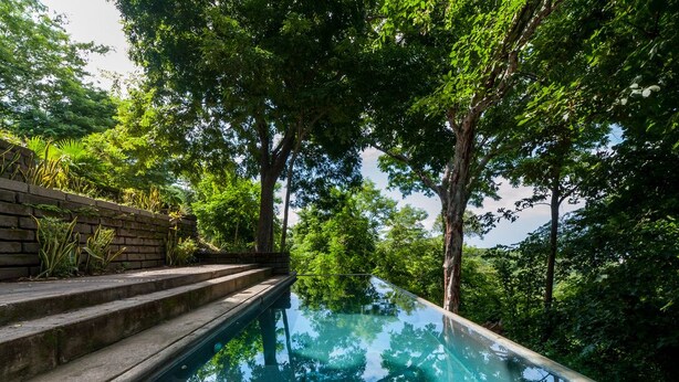 A clear glass infinity pool surrounded by lush greenery and a brick wall. - Jungle Villa with ocean view, shared infinity pool, wildlife, open-air living. (San Juan del Sur)