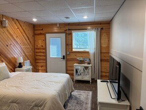 Une chambre à coucher avec un lit, une table de chevet avec une lampe, une fenêtre avec des rideaux, une petite armoire et un téléviseur posé sur un meuble.