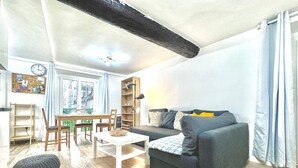 A living room with a dark grey sofa, a white coffee table, and a wooden dining table with chairs. There is a bookshelf, a lamp, and a window with curtains. - Angle droite (Nice)