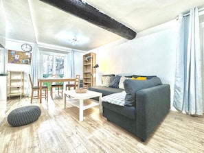 Un salon avec un canapé gris foncé, une table basse blanche et une table à manger en bois avec des chaises. On y trouve également une bibliothèque, une lampe et une fenêtre avec des rideaux.