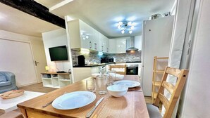 A dining area with a wooden table set for four, a TV in the background, and a kitchen area with modern appliances. - Angle droite (Nice)