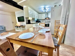 A dining area with a wooden table set for four, a TV in the background, and a kitchen area with modern appliances.