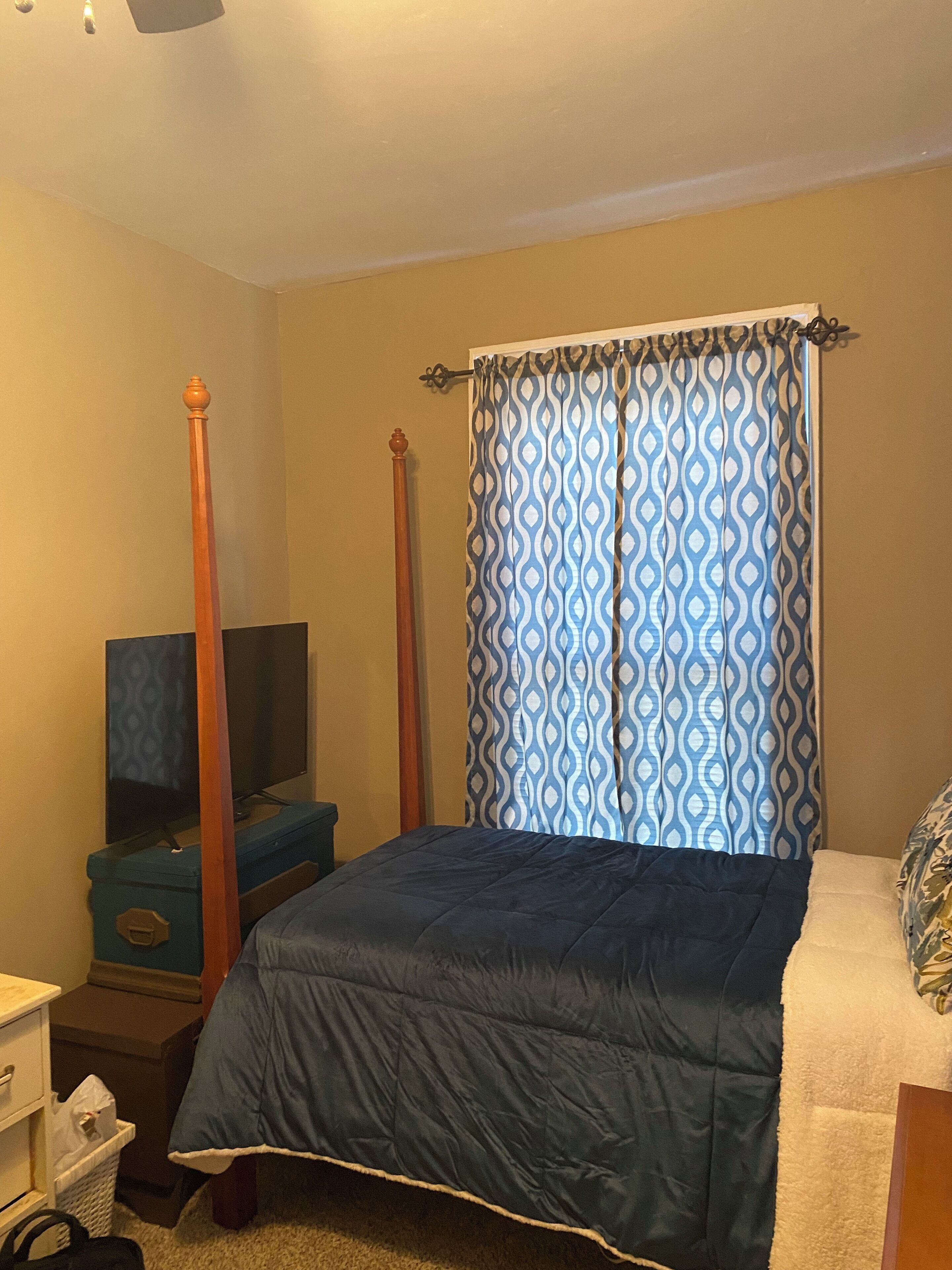 A bedroom with a bed, a television on a stand, a dresser, and a window with patterned curtains.