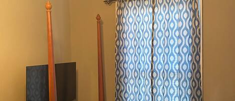 A bedroom with a bed, a television on a stand, a dresser, and a window with patterned curtains.
