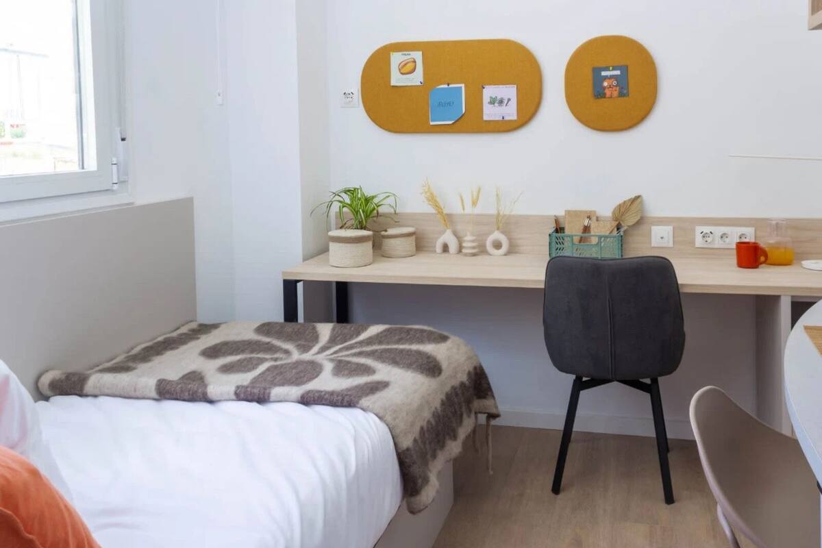 A modern hotel room with a bed, a desk, and a chair. There are bulletin boards on the wall and a window providing natural light.