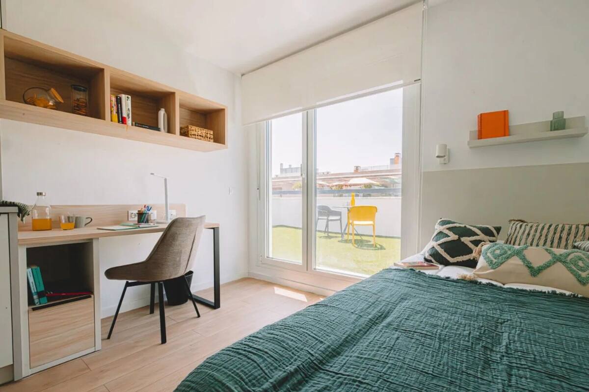 A modern bedroom with a bed, a desk, and a chair. There is a large window providing a view of the outdoors.