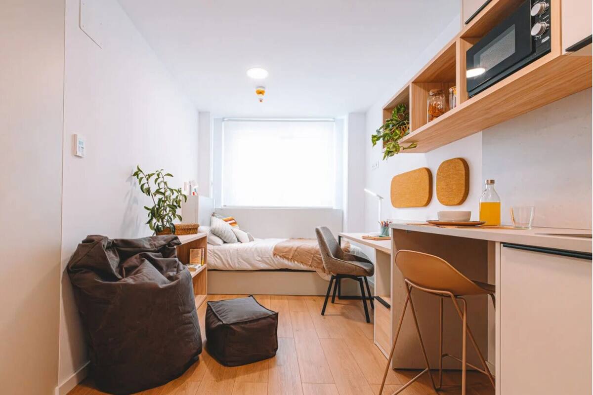 A compact, modern room with a bed, a desk, a chair, a bean bag, and a small kitchenette.