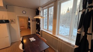 Une pièce comprenant une table à manger, des chaises, un radiateur, une fenêtre donnant sur un paysage enneigé, une horloge au mur et un porte-manteau.