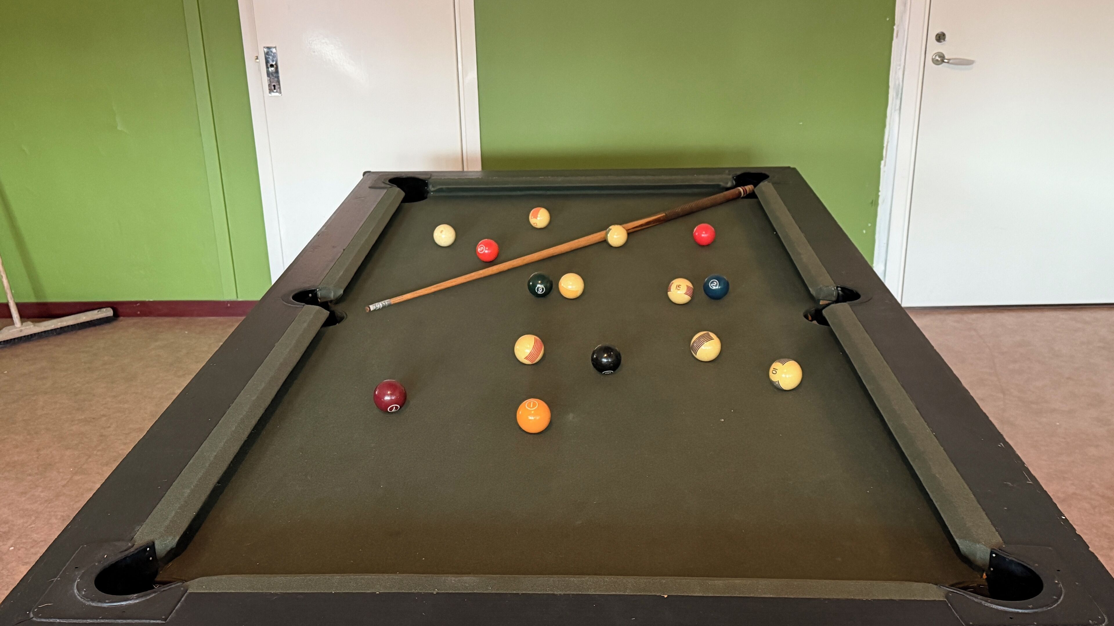 A pool table with balls and a cue stick in a room with green walls and a door.