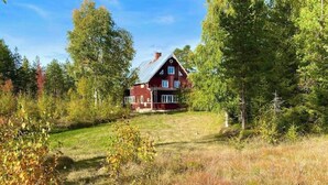 Une maison en bois rouge, avec un toit en pente, entourée d’arbres et d’une zone herbeuse.