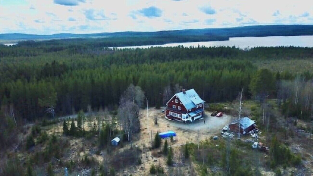 A red cabin with a porch, surrounded by dense forest and a lake in the background.