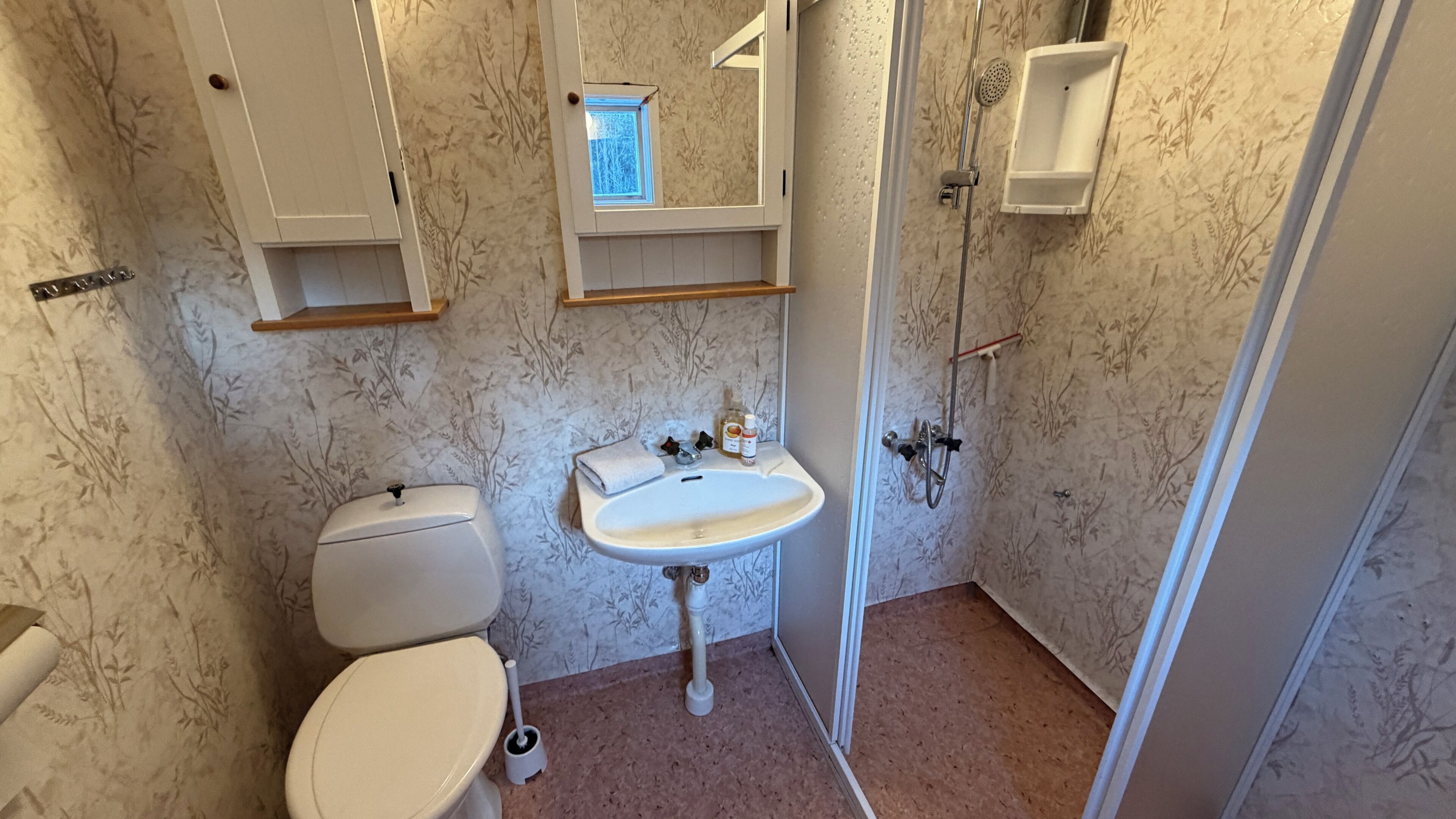 A bathroom with a toilet, sink, and shower. There are shelves above the sink and a window above the toilet.