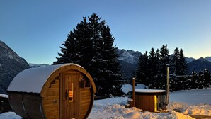 A wooden cabin with a snow-covered roof, a sauna, and a view of snow-covered mountains. - Bergbijou - Luxurious vacation home with sauna, whirlpool, infrared and mountain views (Scuol)