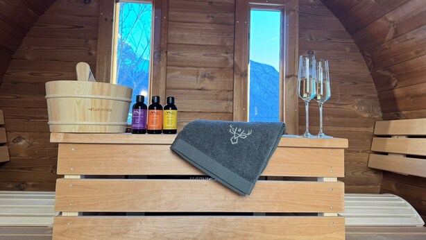 A wooden cabin interior with a bench, a wooden bucket, three bottles, a towel, and two glasses on the bench. - Bergbijou - Luxurious vacation home with sauna, whirlpool, infrared and mountain views (Scuol)