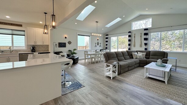 A spacious living area with a large gray sectional sofa, a dining table, and a kitchen with white cabinets and pendant lighting. - True Luxury Waterfront: Dock, Kayaks & Hot Tub (Ocean Shores)