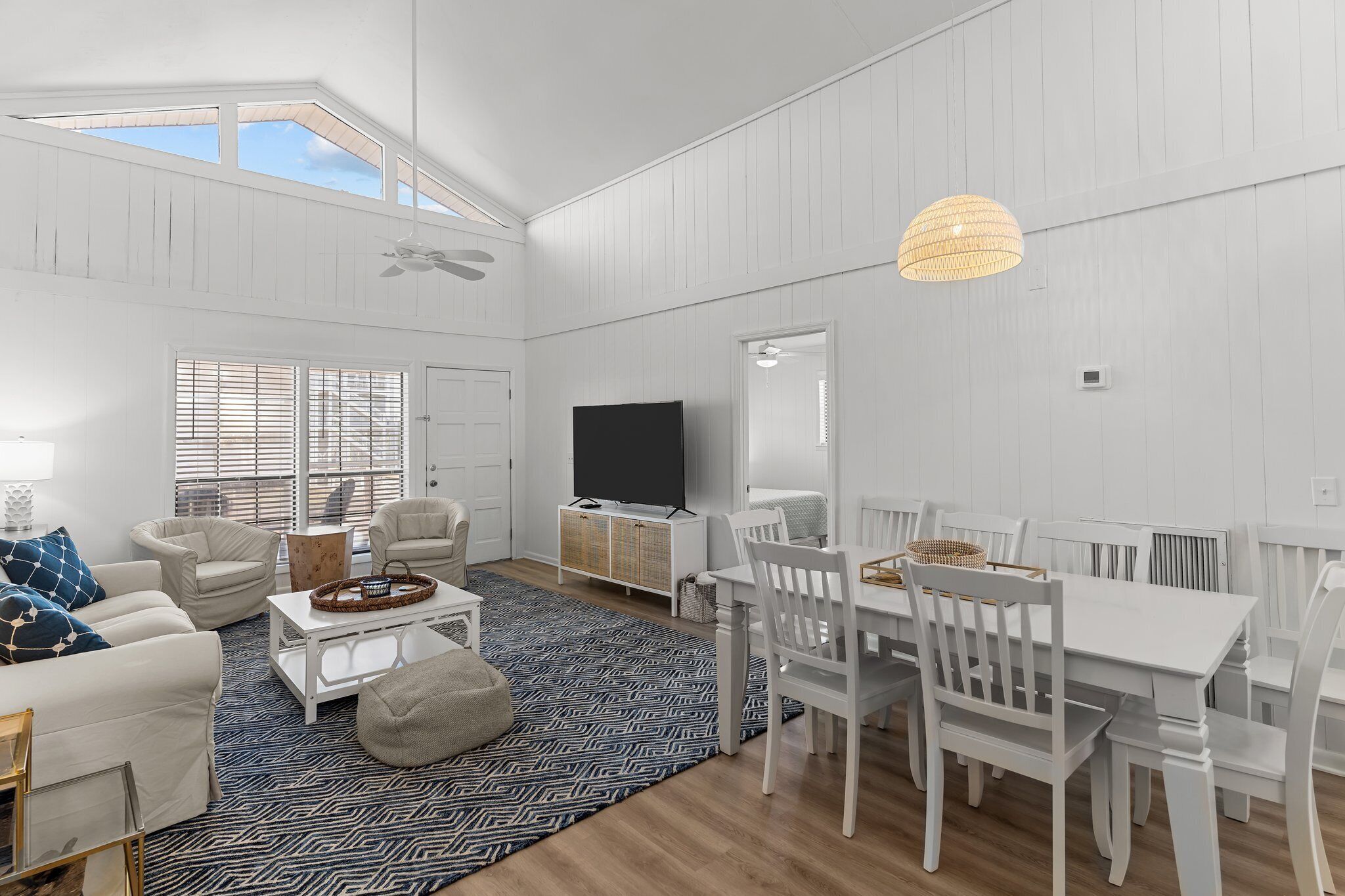 A spacious living room with a high ceiling, a large TV, white furniture, and a patterned rug.