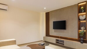 Una sala moderna con un televisor de pantalla plana montado en la pared, una mesa redonda de centro, un sofá color beige y una estantería decorativa con plantas.