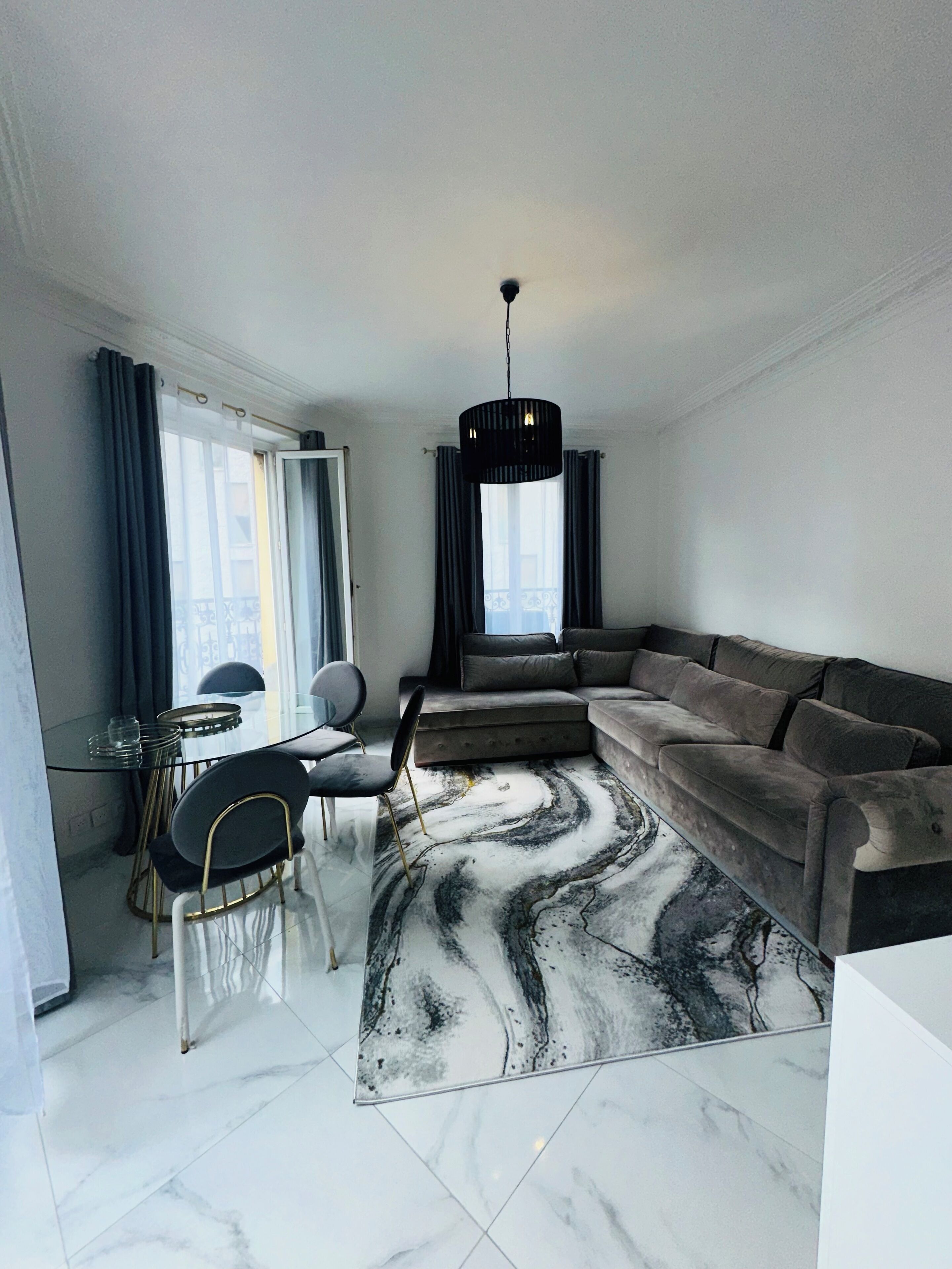 Een moderne woonkamer met een hoekbank, een eethoek met een glazen tafel en een groot raam met gordijnen.