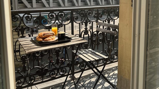 Un balcón con una mesa dispuesta para el desayuno, con cruasanes y jugo de naranja, con vistas a un paisaje urbano que incluye edificios y un puente.