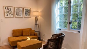 Un salon comprenant un canapé, un pouf et un fauteuil avec un coussin à motif léopard. Trois œuvres d’art encadrées ornent le mur, ainsi qu’une lampe sur pied et une fenêtre donnant sur un paysage verdoyant.