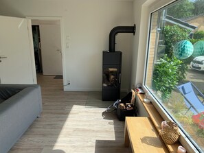 Un espacio habitable compacto con sofá, una pequeña mesa de madera, un calentador negro y una ventana que ofrece vistas a un coche y zonas verdes.