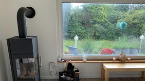 A room with a black wood stove, a window with a view of a garden, a wooden bench, and a small table with decorative items.
