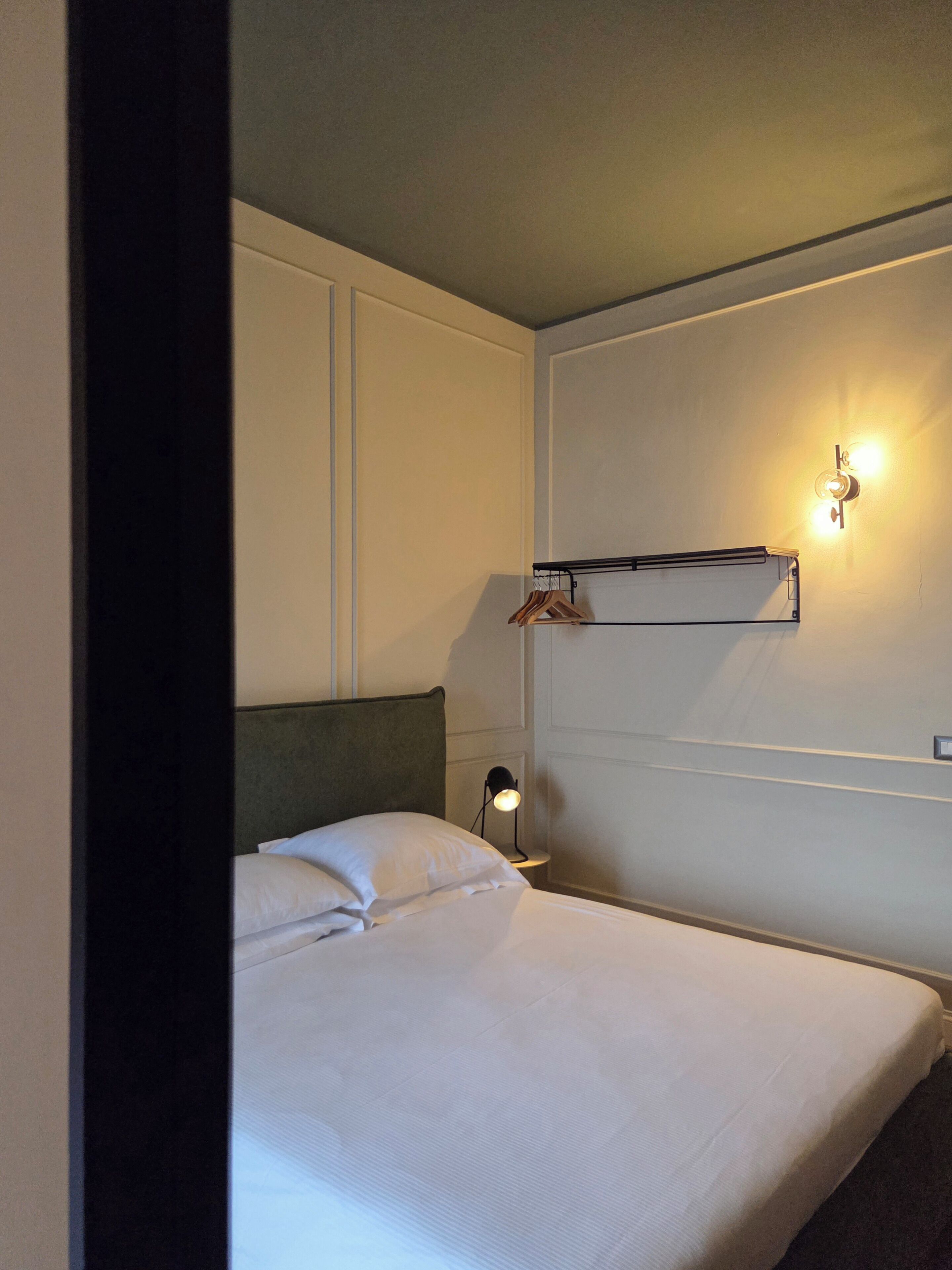 A hotel room with a bed, a wall-mounted shelf with clothes, a nightstand with a lamp, and a wall-mounted light fixture.
