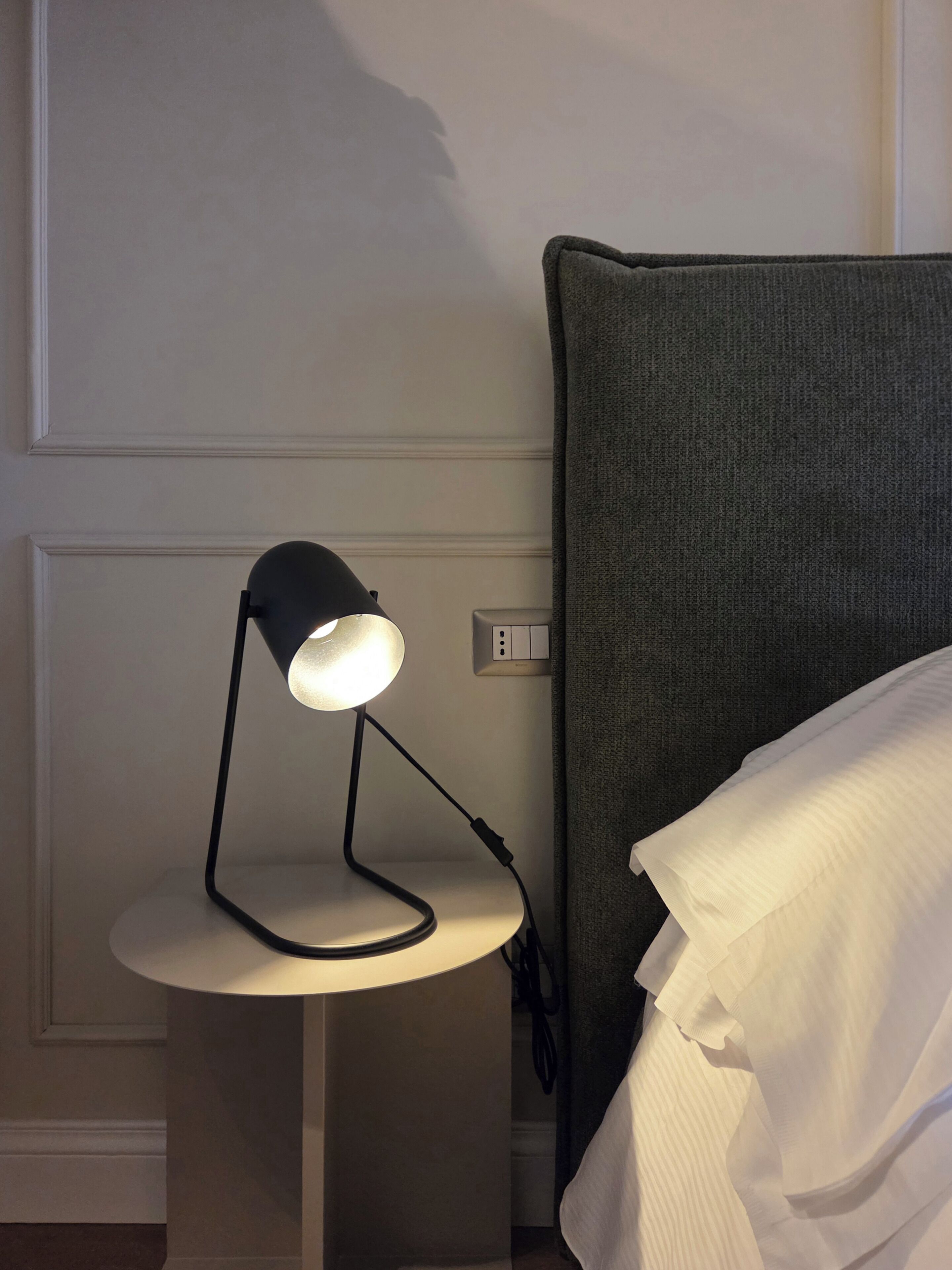 Uma mesa de cabeceira com uma luminária, uma tomada e uma cama com cabeceira cinza.