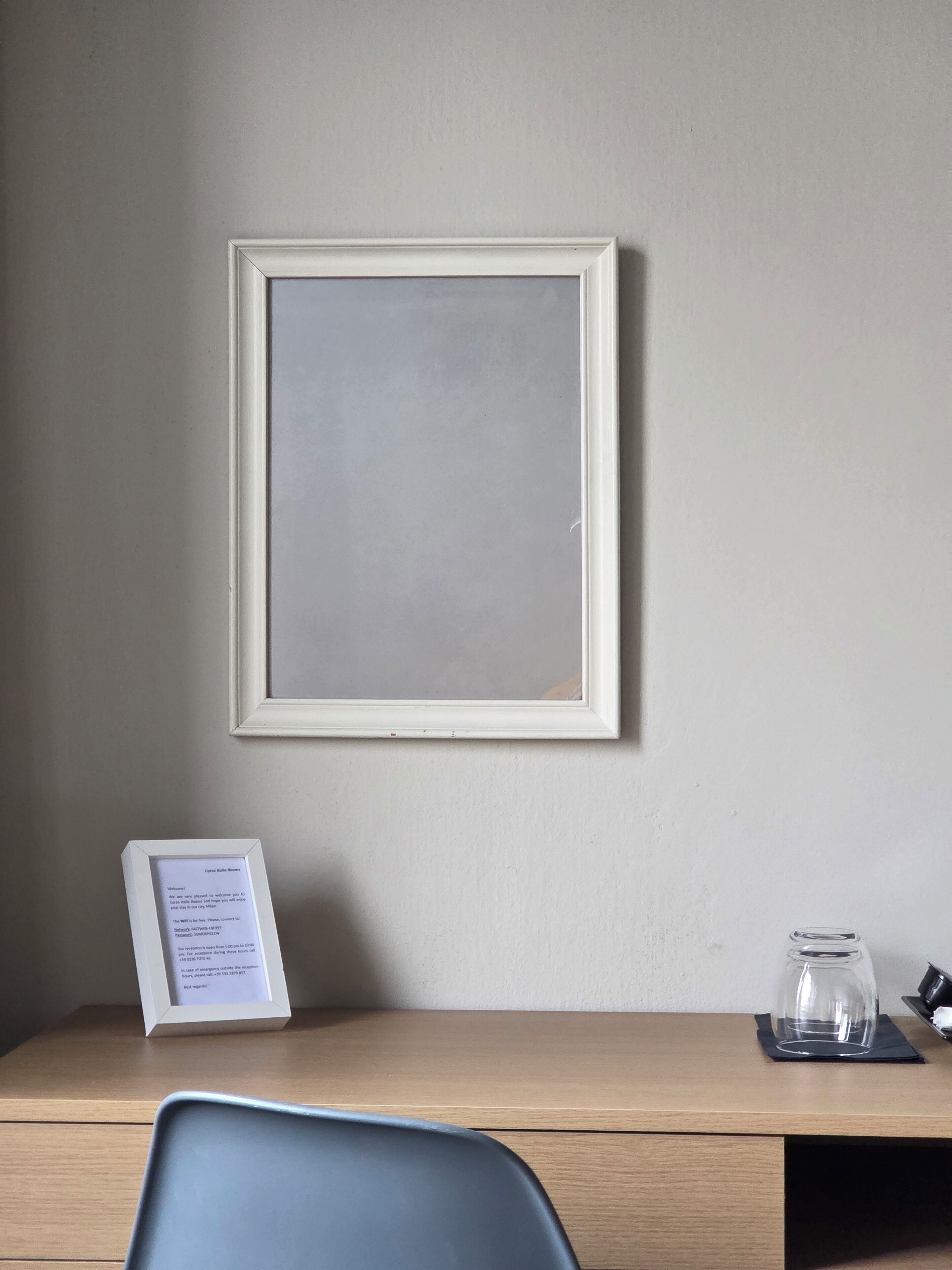 A hotel room with a wooden desk, a framed empty picture, a glass jar, and a welcome sign.