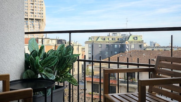 A balcony with a view of buildings, a potted plant, and wooden furniture. - Corso Italia Rooms (Milan)