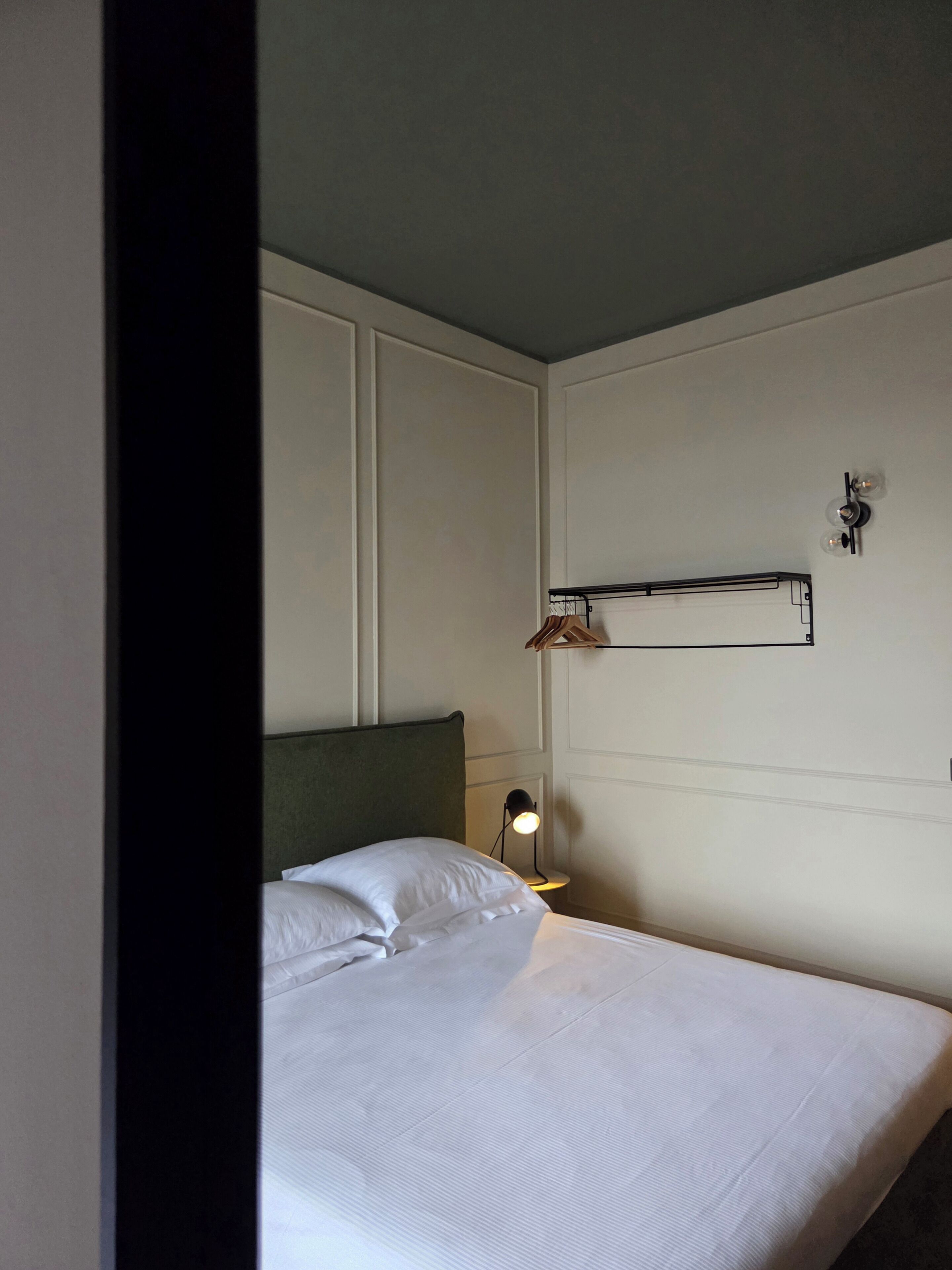 Habitación de hotel con cama, lámpara de noche y estante en la pared con ropa colgada.