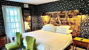 A neatly made bed with pillows, a green armchair, a wooden nightstand, and a window with curtains. - The Bond Inn Room #5 (Warrensburg)