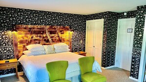 A bedroom with a bed, two green chairs, a wooden headboard, a white door, and floral wallpaper. - The Bond Inn Room #5 (Warrensburg)