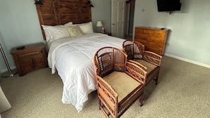 A bedroom with a large bed, two wicker chairs, a wooden dresser, a nightstand, and a wall-mounted TV. - The Bond Inn Room #7 (Warrensburg)