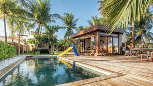 Un área de piscina con terraza de madera, un tobogán y una zona de estar cubierta con mesas y sillas.