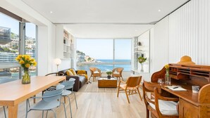 A modern living room with a large window overlooking a beach, a dining area with a wooden table, and a seating area with a sofa and chairs. - Apartment Seacliff - Two Bedroom Apartment, Sleeps 4 (Cape Town)
