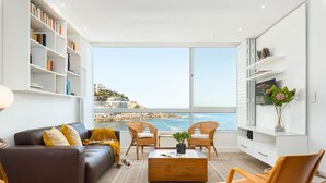 A modern living room with a large window overlooking a coastal view, featuring a sofa, chairs, a coffee table, and a bookshelf. - Apartment Seacliff - Two Bedroom Apartment, Sleeps 4 (Cape Town)