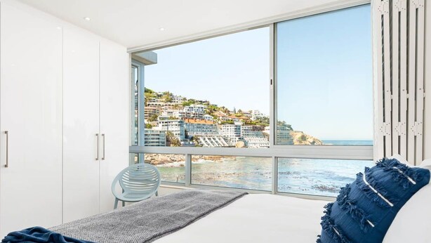 A bedroom with a large window overlooking a coastal view, white furniture, and a view of buildings on a hillside. - Apartment Seacliff - Two Bedroom Apartment, Sleeps 4 (Cape Town)