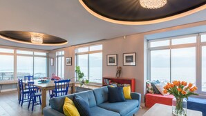 Ein modernes Wohnzimmer mit einem blauen Sofa, einem Esstisch mit Stühlen und einem großen Fenster mit Blick aufs Meer.