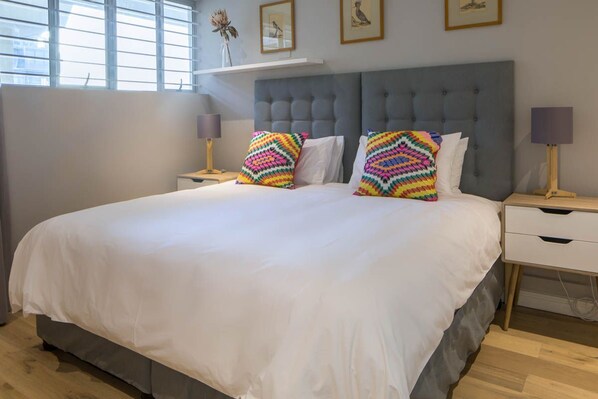A large bed with white bedding and colorful pillows, a grey upholstered headboard, and bedside tables with lamps.